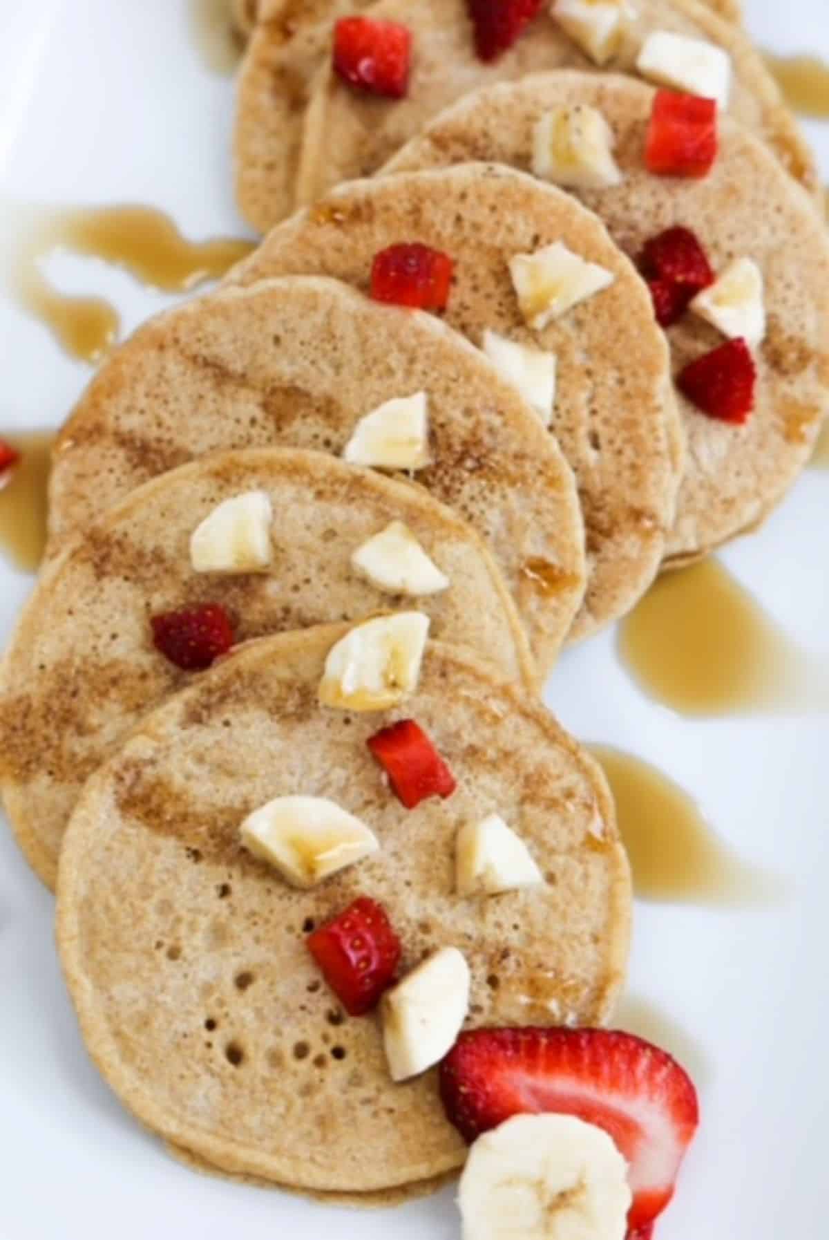 Several small pancakes overlapping, topped with pieces of banana and strawberry. Drizzled with maple syrup.