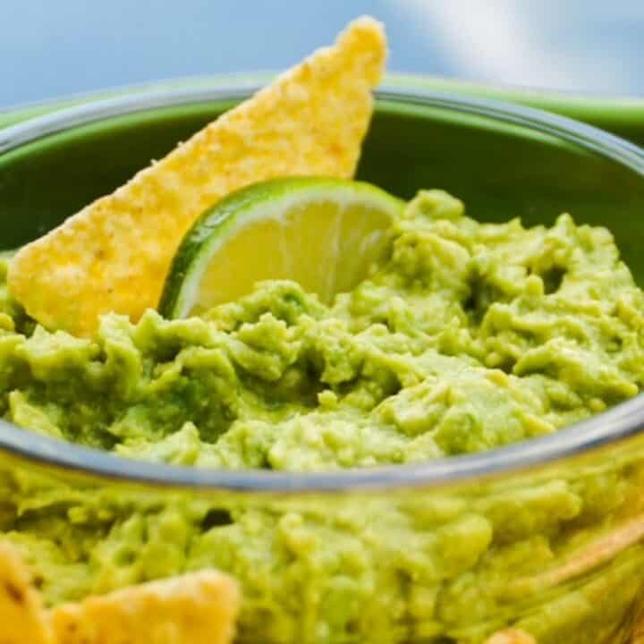 Guacamole in a glass bowl with one chip and a slice of lime.
