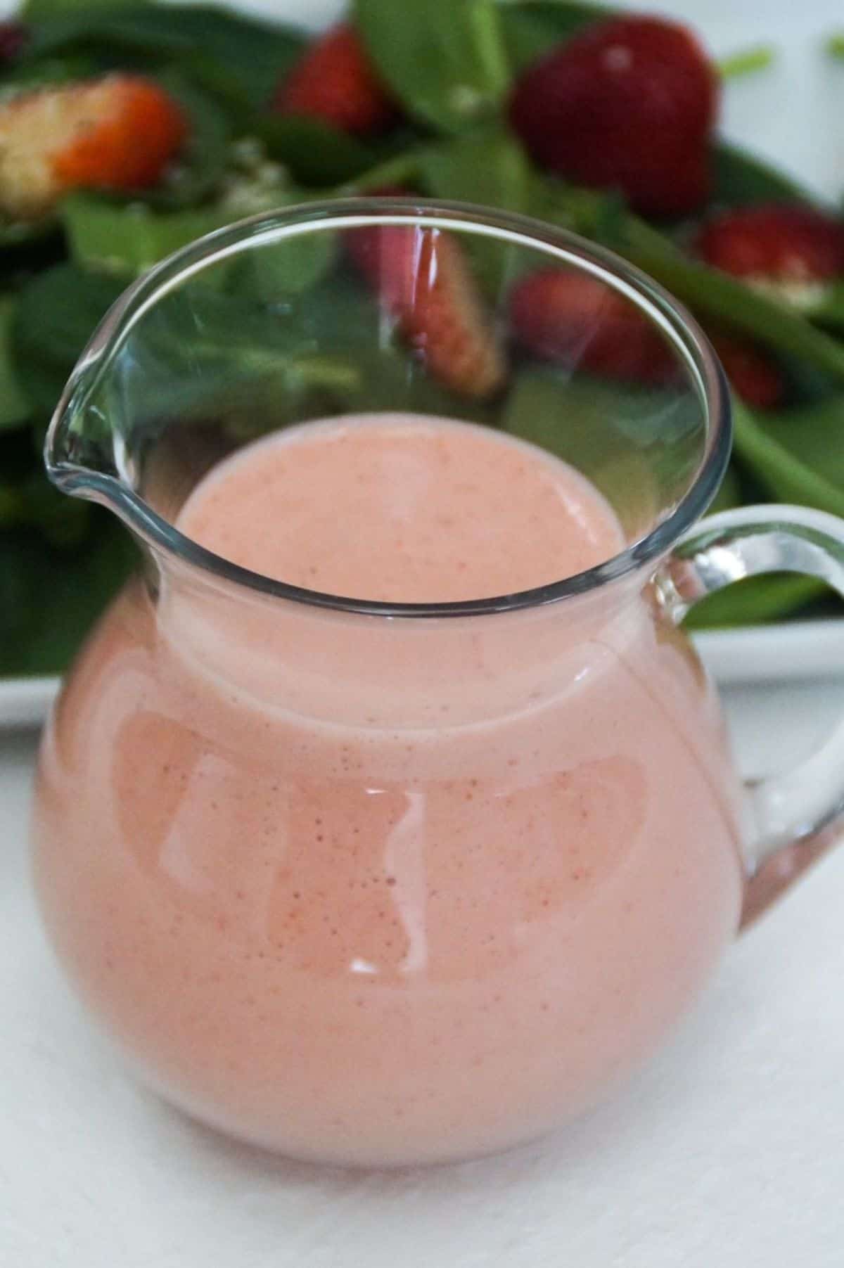 Strawberry salad dressing in a glass carafe with spinach salad behind it.