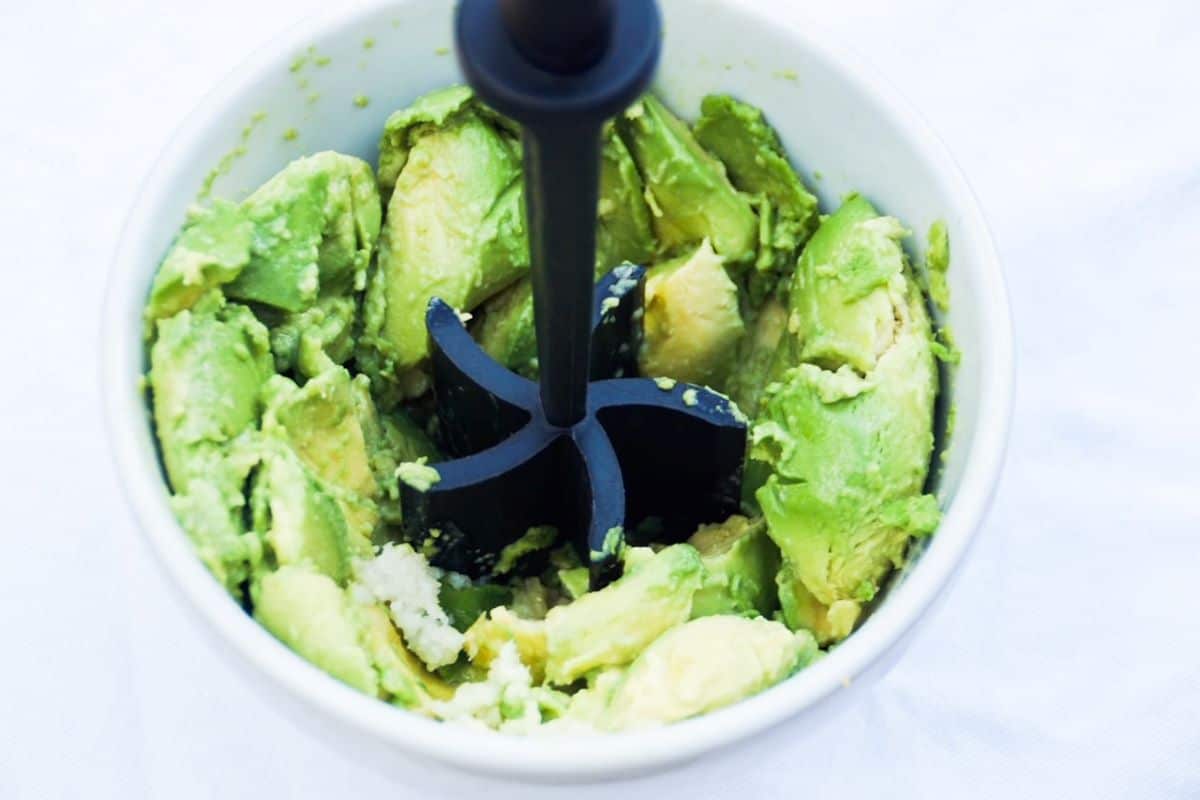 Guacamole ingredients being pressed with a food masher in a white bowl.