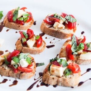 Six pieces of caprese bruschetta drizzled with balsamic glaze on a white platter.