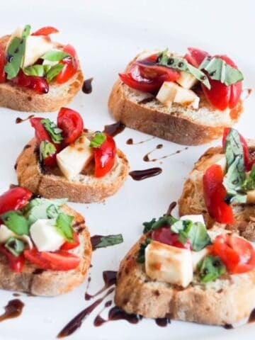 Six pieces of caprese bruschetta drizzled with balsamic glaze on a white platter.