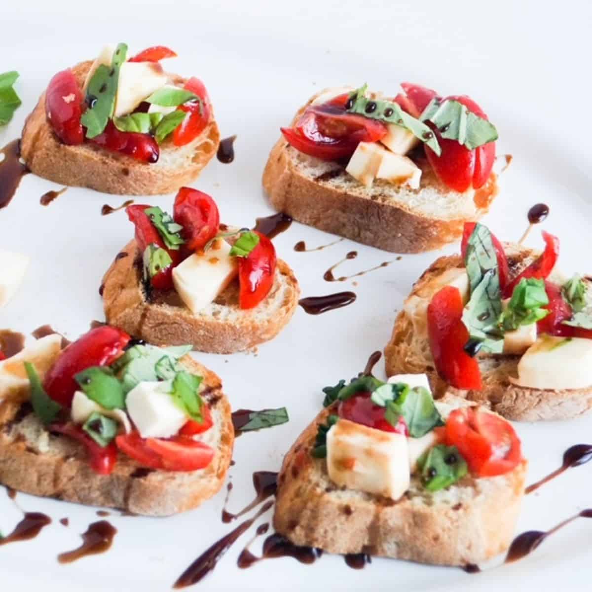 Six pieces of caprese bruschetta drizzled with balsamic glaze on a white platter. 