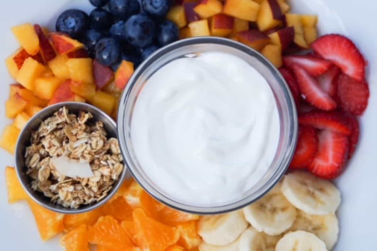 Greek yogurt in a glass bowl with a small bowl of granola centered in the middle of a variety of fruit (nectarine pieces, blueberries, sliced strawberries, sliced banana, and orange pieces).