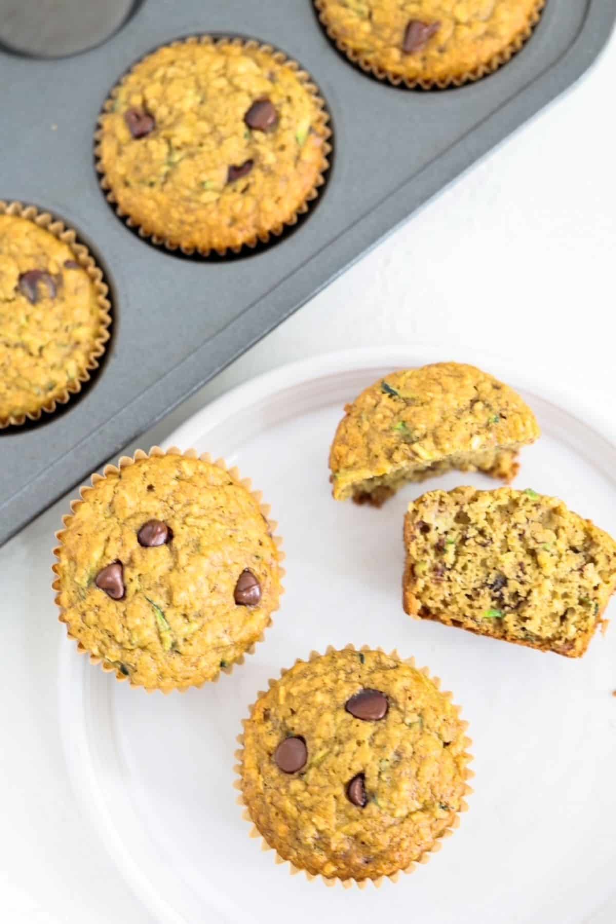 Two whole and two halves of banana chocolate chip muffins on a whit plate with a muffin tin of muffins behind it.