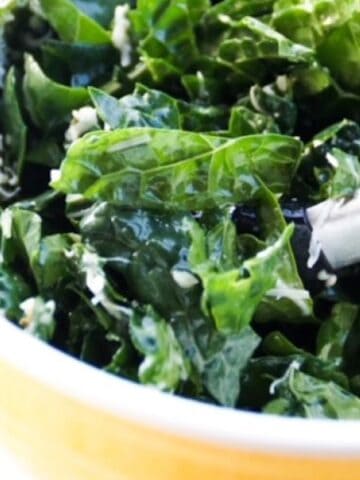 Kale salad in a yellow bowl with a set of tongs in the salad as to serve.