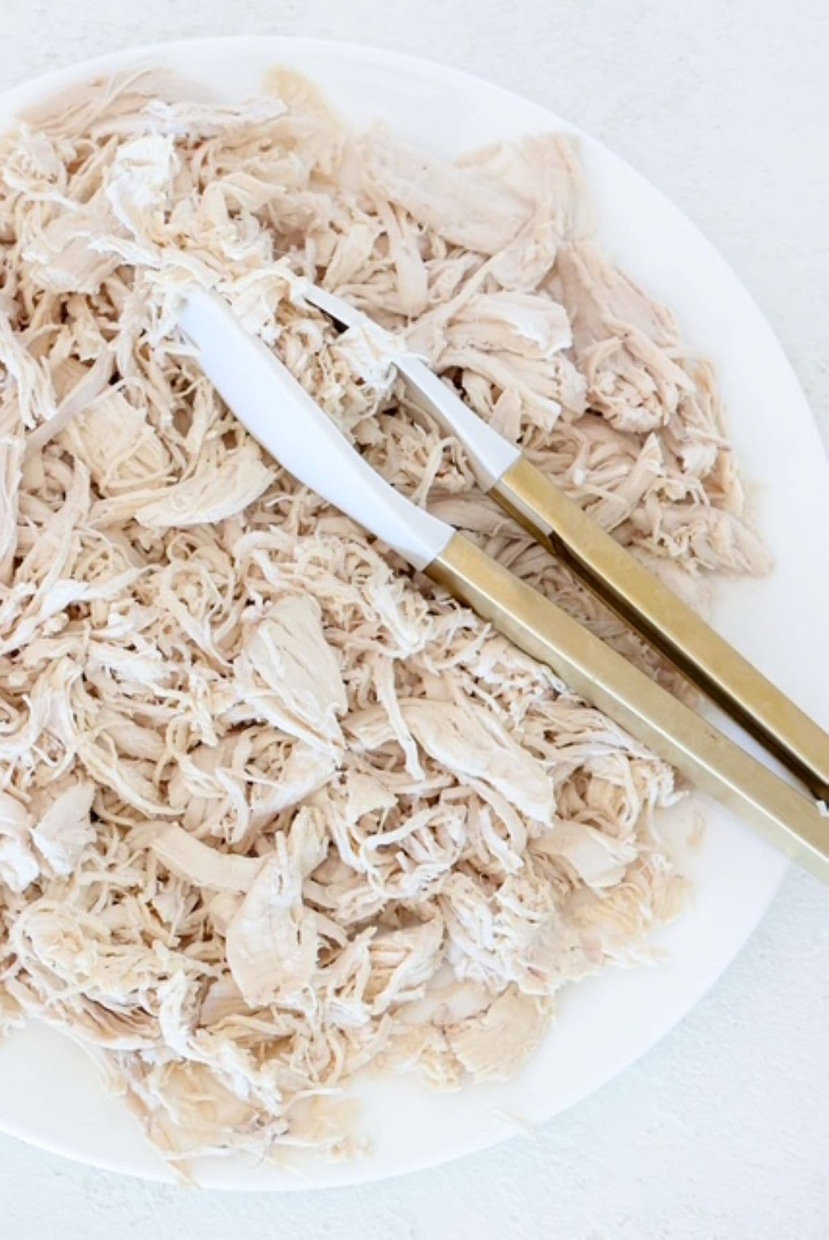 Shredded chicken on a white plate with white and gold serving tongs.