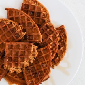 Quarters of round pumpkin waffles overlapping and topped with a drizzle of maple syrup on a white plate.