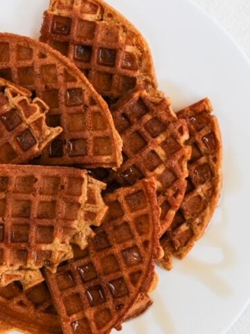 Quarters of round pumpkin waffles overlapping and topped with a drizzle of maple syrup on a white plate.