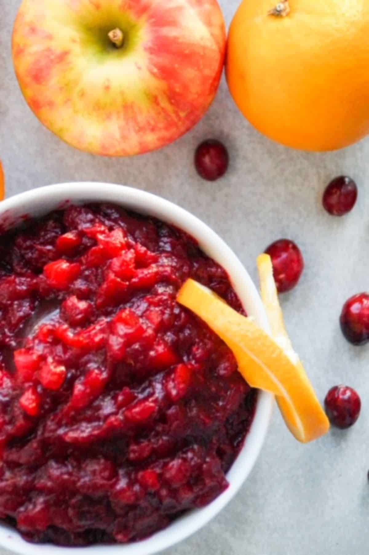 Cranberry sauce in a white bowl with an orange slice wrapped around the edge on the right. One apple and one orange are above the bowl with a few cranberries scattered between.