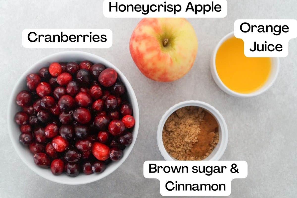 Cranberry sauce in a white bowl to the left, labeled. One Honeycrisp apple, orange juice in a small white prep bowl, and brown sugar with cinnamon in a separate white prep bowl to the right of the cranberry sauce. Each dish is labeled.