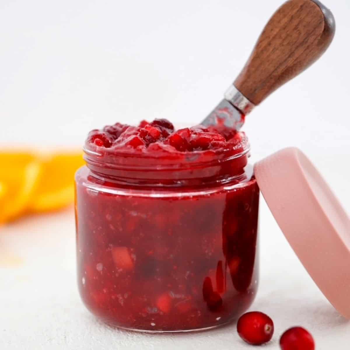 Homemade low sugar cranberry sauce in a glass jar, with the lid and a few cranberries to the side.