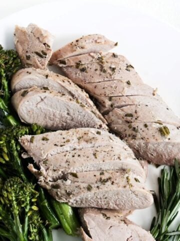 Slices of roasted pork tenderloin with broccolini to the left and fresh rosemary to the right of the cooked tenderloin.