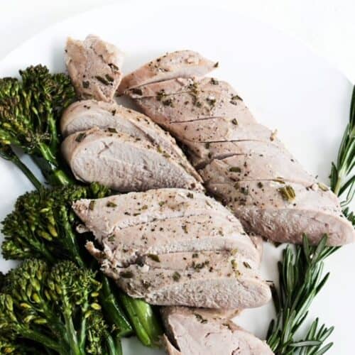 Slices of roasted pork tenderloin with broccolini to the left and fresh rosemary to the right of the cooked tenderloin.