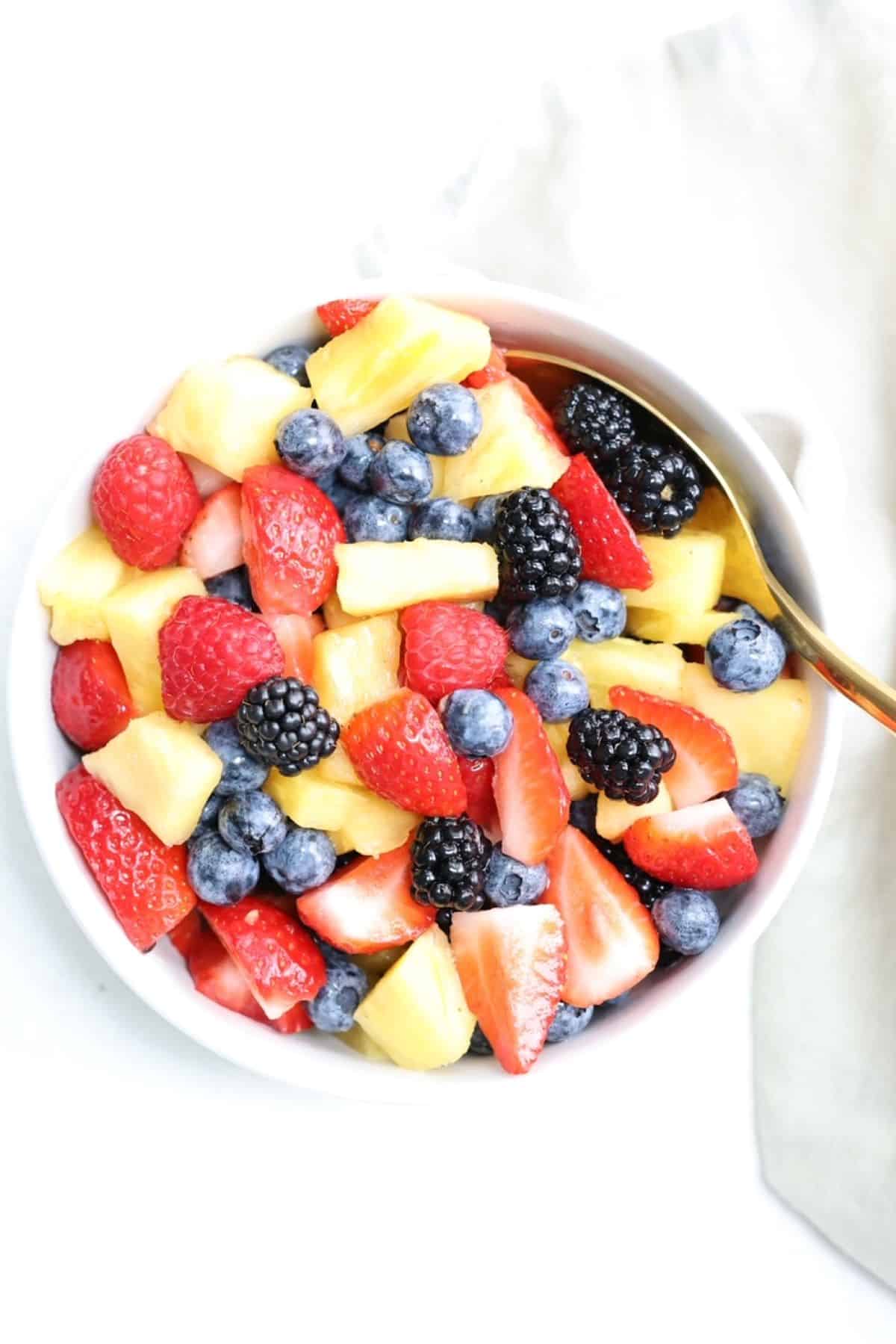 Top view of a bowl of fresh fruit salad with a gold spoon and linen napkin to the side.