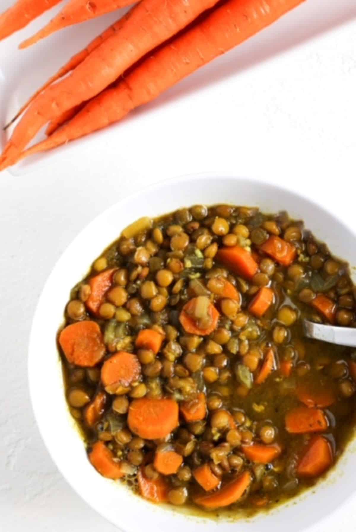 Carrot and Lentil Soup in a white bowl with a white spoon with one bunch of carrots behind the bowl of soup.