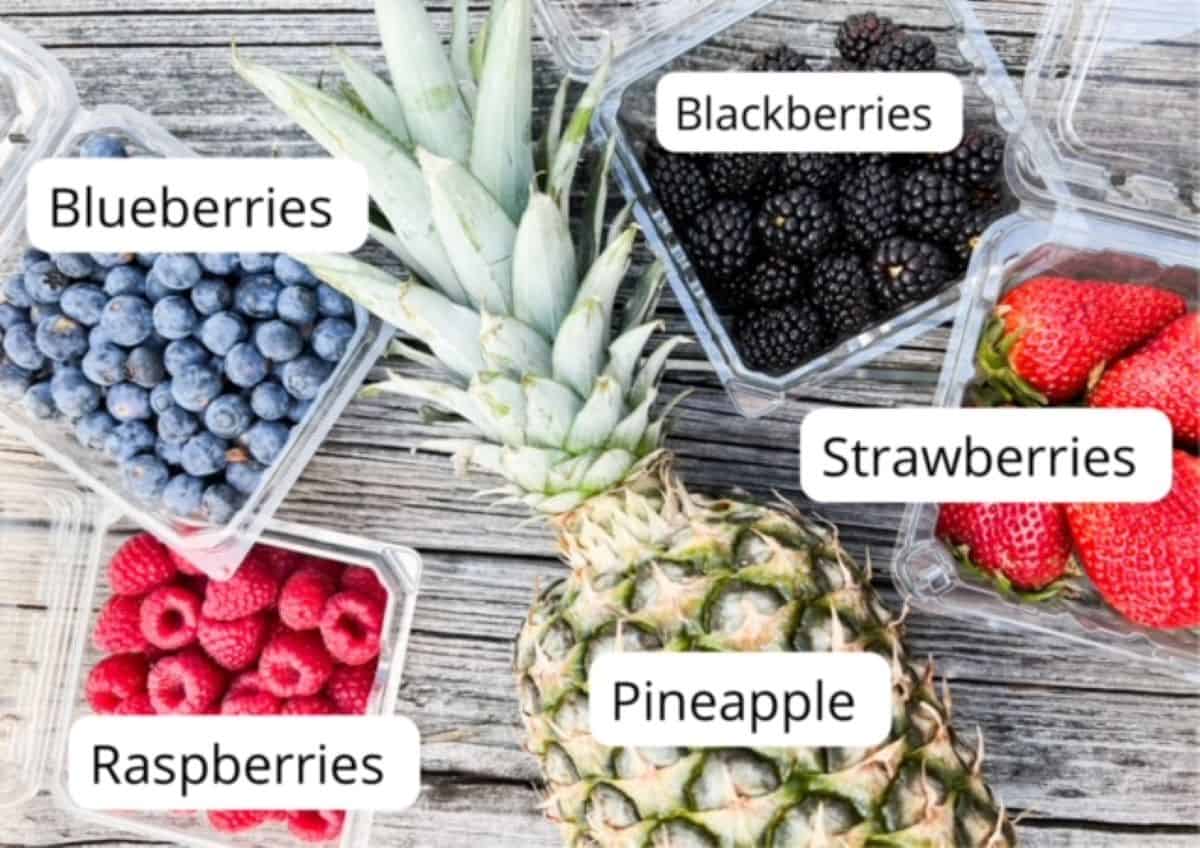 One whole pineapple surrounded by individual containers of raspberries, blueberries, blackberries, and strawberries as you would buy from the grocery store.