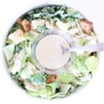 One bowl of Caesar salad with a small glass bowl of Caesar dressing with a spoon in the center.