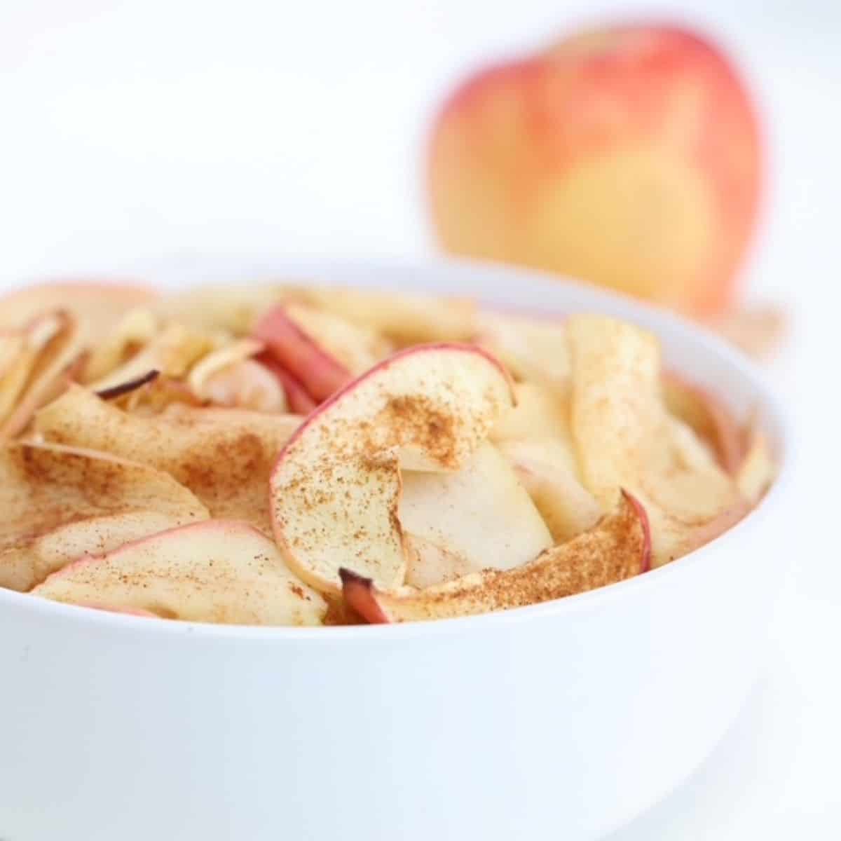 Air fried apple slices in a white bowl with an apple in the background.