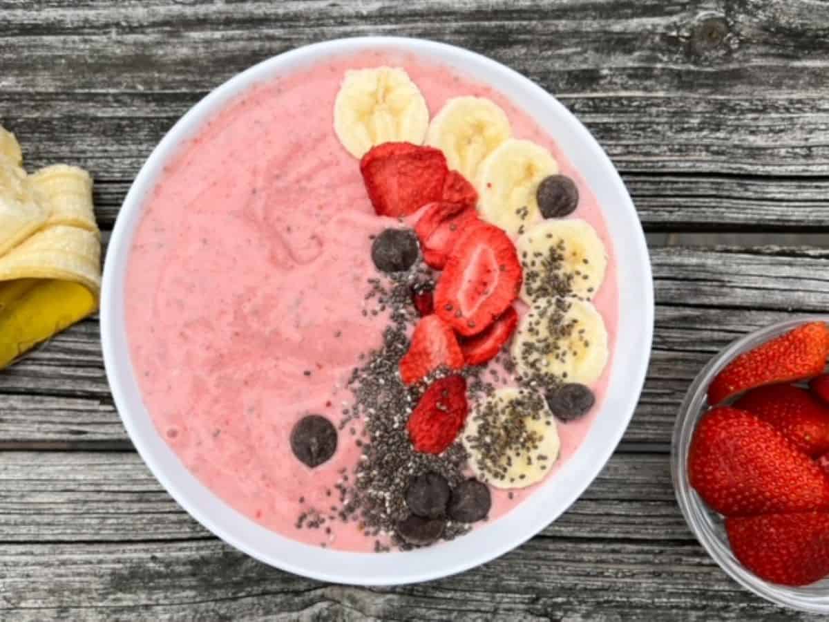 Strawberry banana smoothie bowl with toppings served in a white bowl with a half of a banana to the left and strawberries to the right of the bowl.