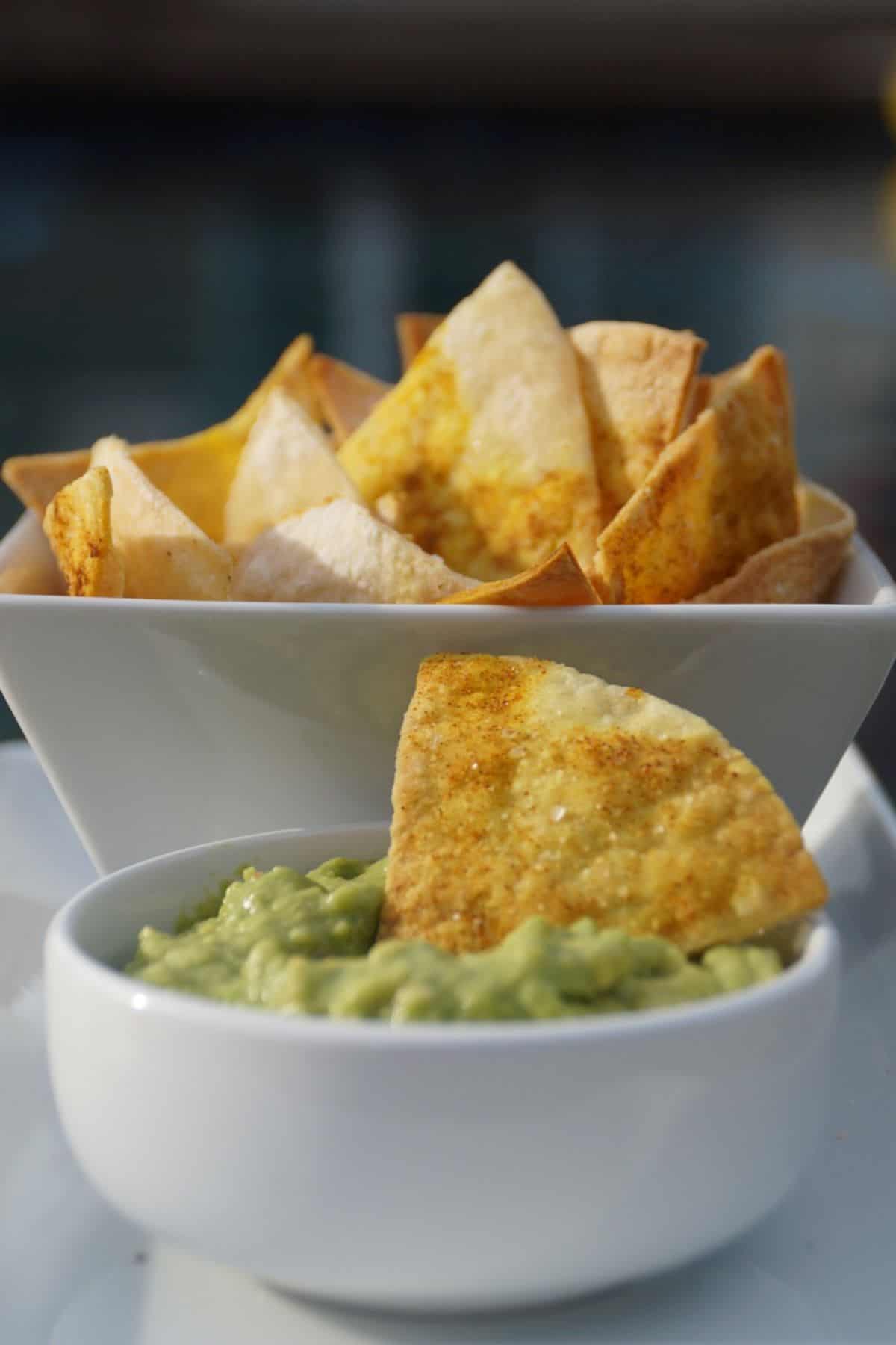 Tortilla chip in a bowl of guacamole with a bowl of chips in the background.