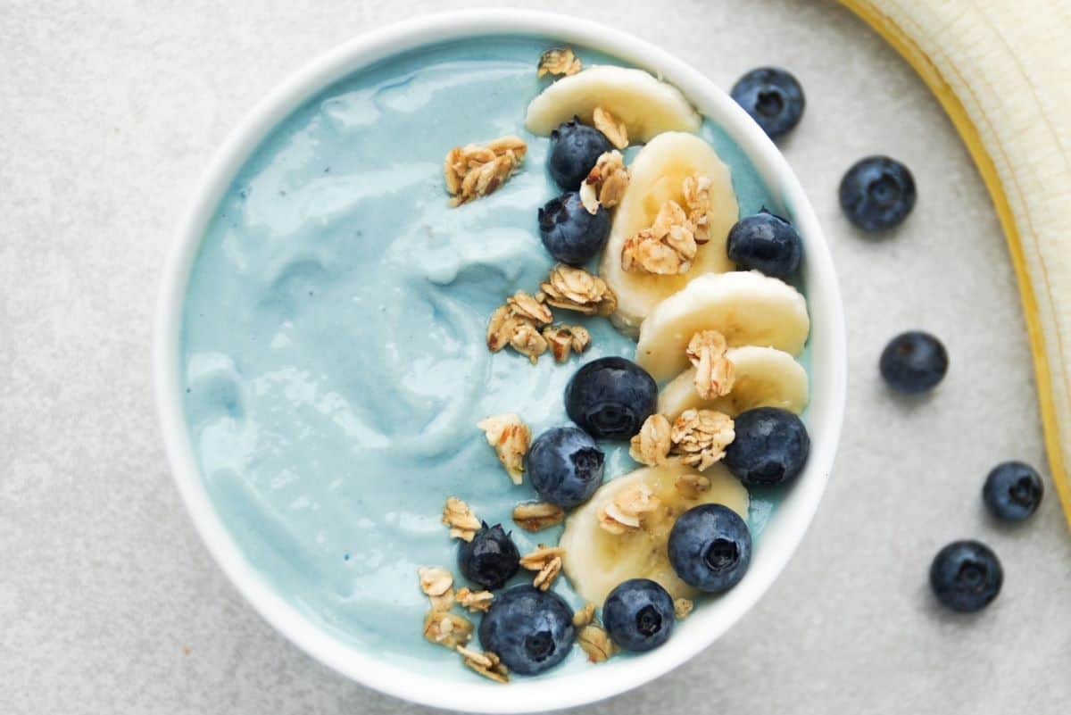 Blue smoothie bowl served in a white bowl, topped with blueberries, banana slices, and granola. Blueberries and a peeled banana are to the right of the bowl.