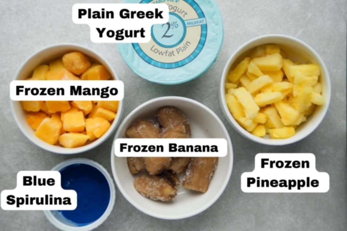 One container of 2% low fat Greek yogurt and four white bowls, each one having one ingredient: frozen mango, frozen banana pieces, frozen pineapple, and one with blue spirulina. Each ingredient is labeled.