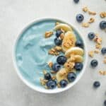 Blue smoothie bowl served in a white bowl and topped with granola, banana slices, and blueberries.
