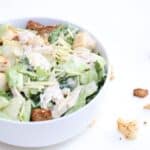 One chicken Caesar salad in a white bowl with croutons to the right of the bowl.