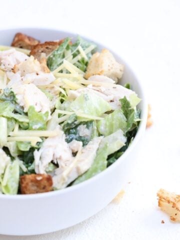 One chicken Caesar salad in a white bowl with croutons to the right of the bowl.