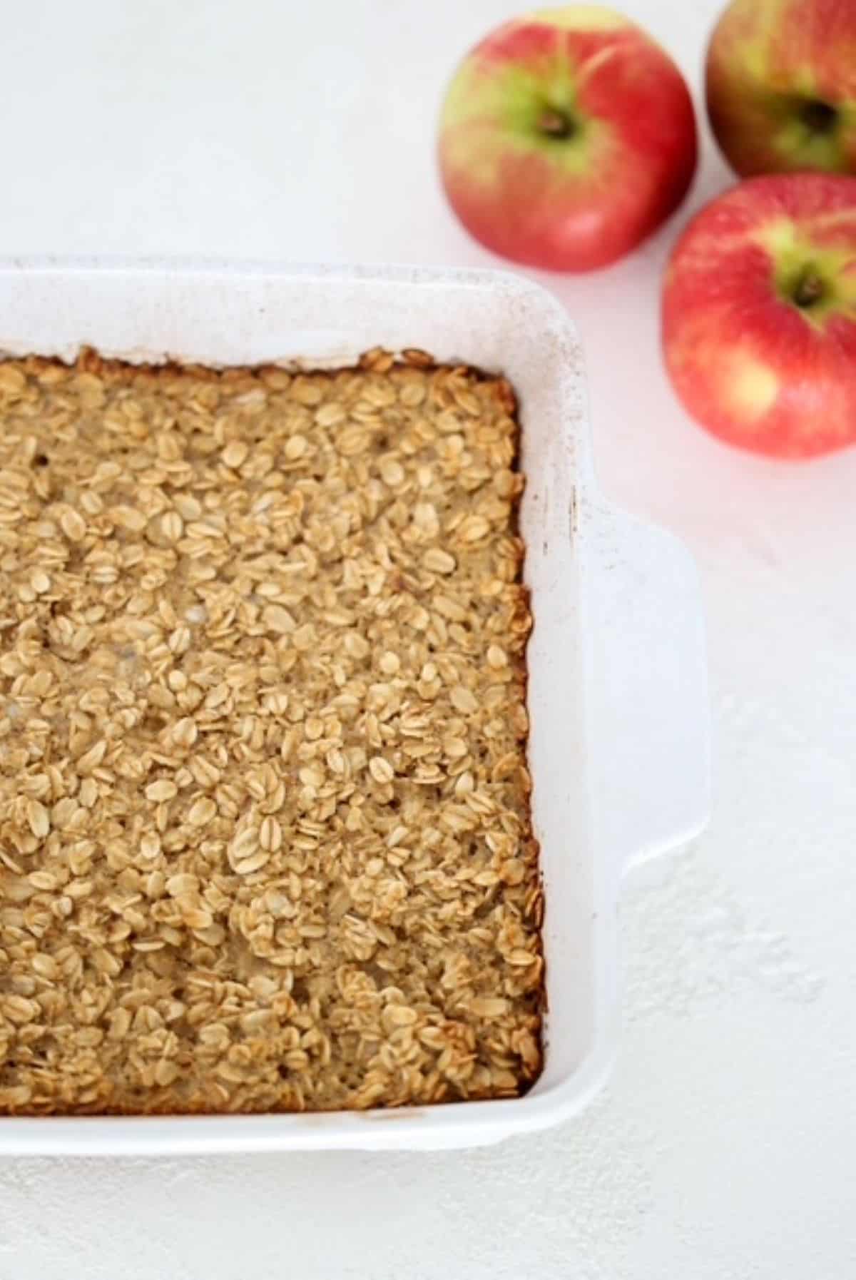 Apple Cinnamon Baked Oatmeal in a white baking dish with three apples to the top right of the baked oatmeal.