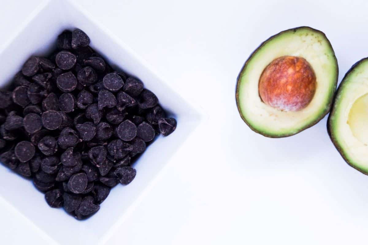 Chocolate chips in a square white bowl and one avocado, halved.