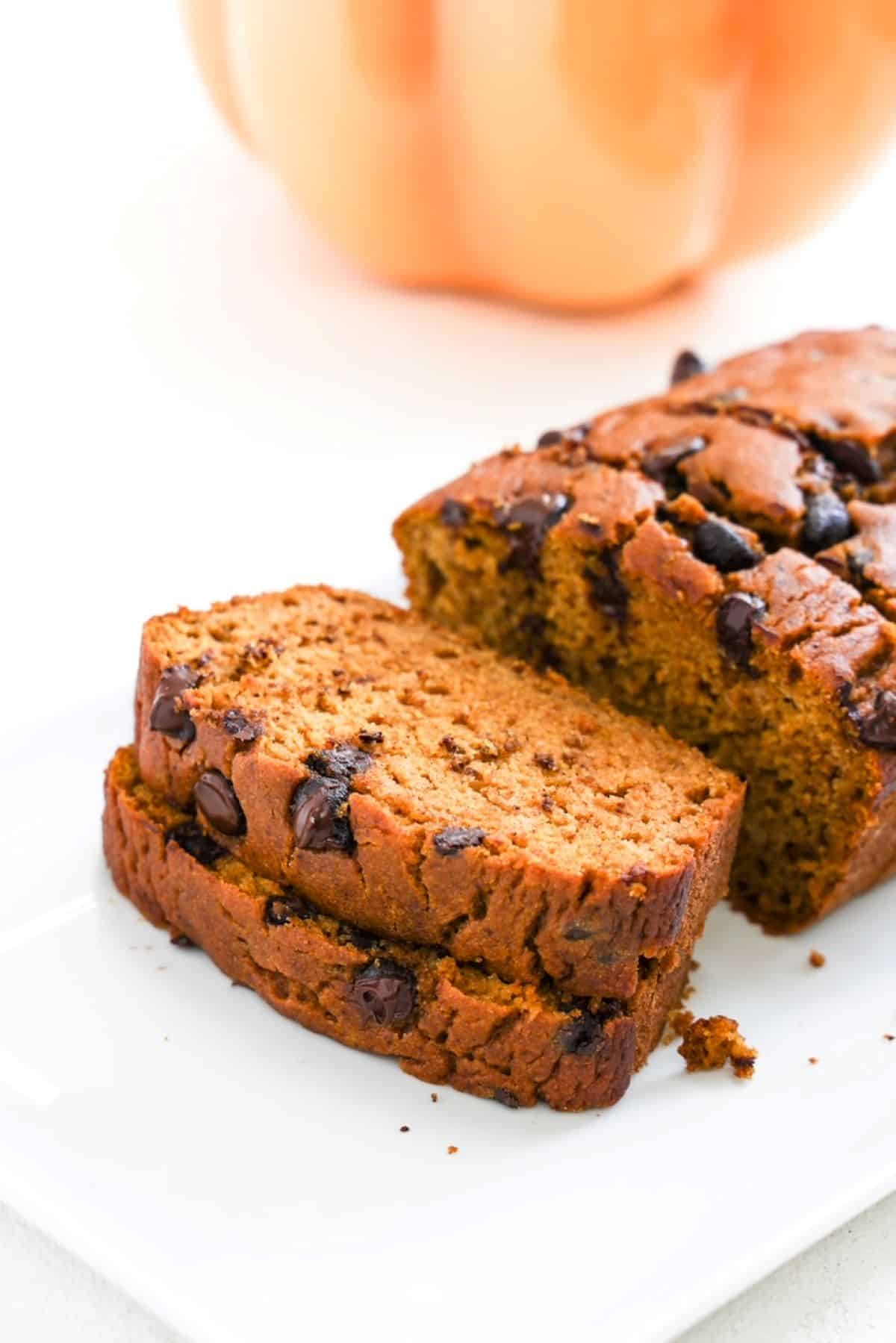 Two slices of pumpkin chocolate chip bread stacked in front of the rest of the loaf. Part of a decorative orange pumpkin is in the background.