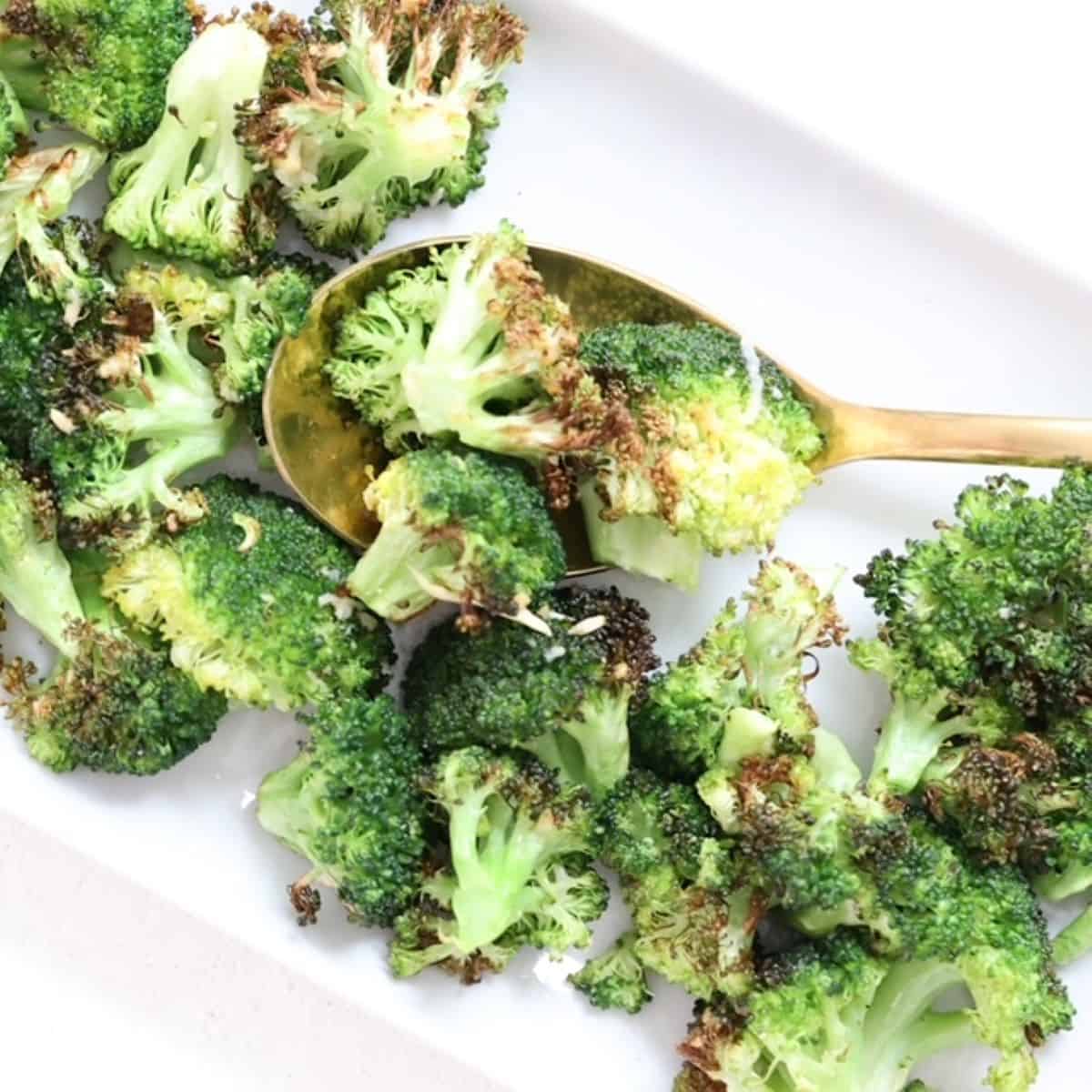 Roasted broccoli on a white rectangular shaped plate with a gold spoon.