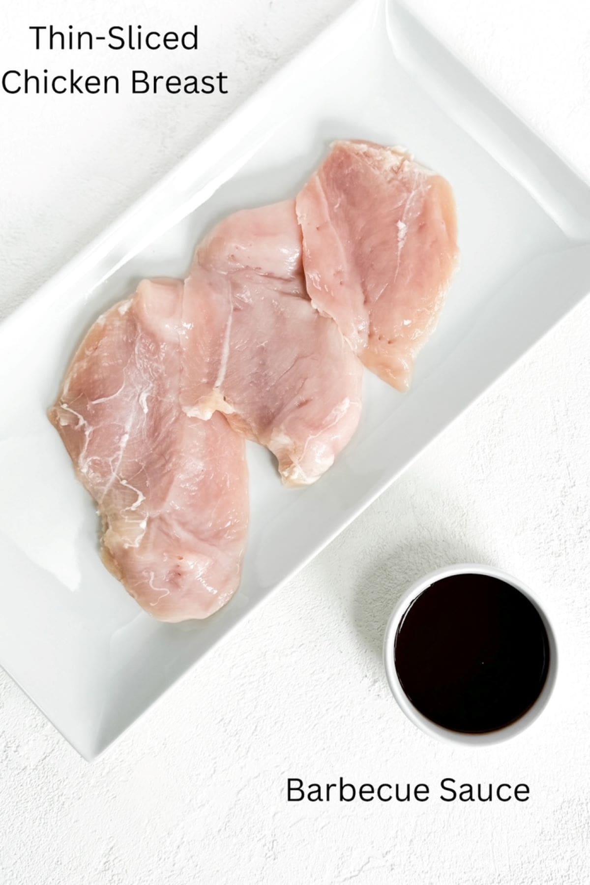 Three pieces of uncooked thin-sliced chicken breasts on a white plate with a small ramekin of barbecue sauce, labeled.