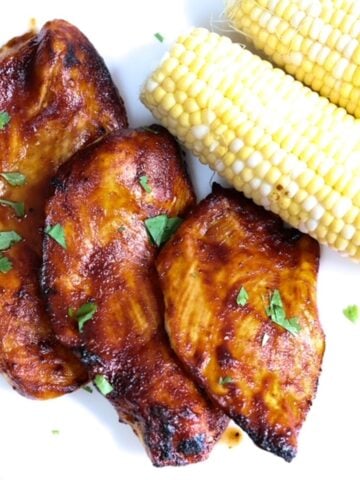 Three pieces of barbecue chicken with two ears of corn topped with pieces of cilantro and served on a white plate.