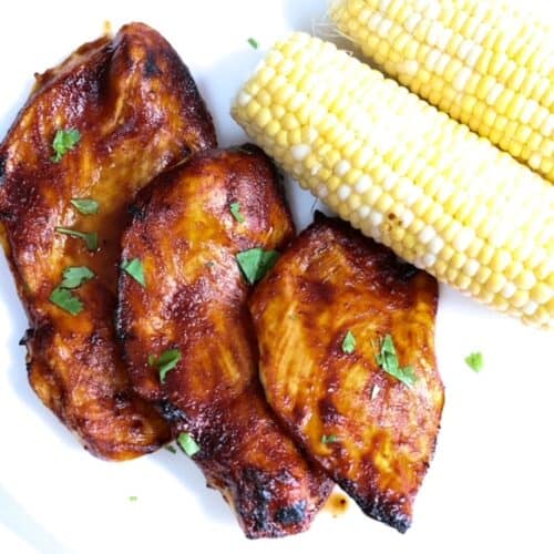 Three pieces of barbecue chicken with two ears of corn topped with pieces of cilantro and served on a white plate.