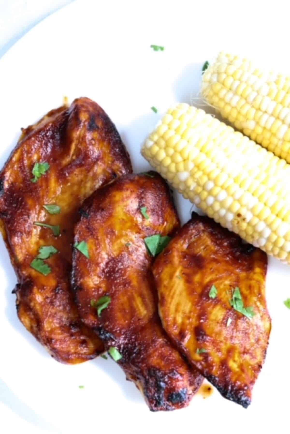 Three pieces of barbecue chicken with two ears of corn on a white plate.