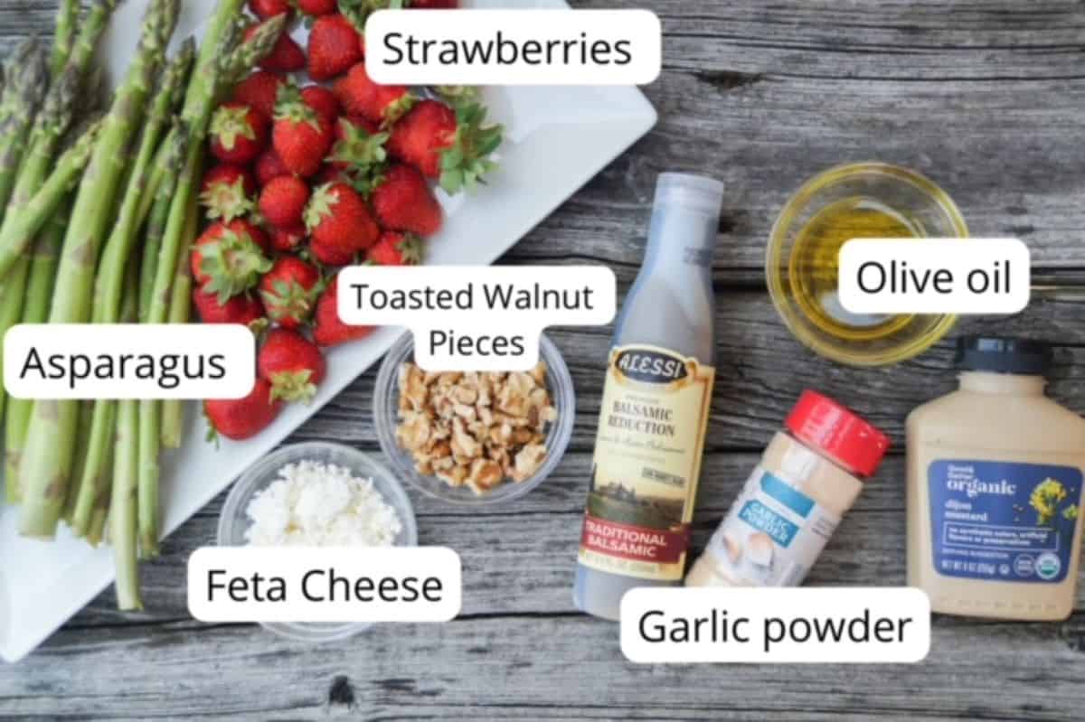 Ingredients for cold asparagus salad with strawberries on a piece of wood and labeled: asparagus and strawberries on a white plate. Feta cheese, walnuts, and olive oil are in separate small glass bowls. Garlic powder container and Dijon Mustard container.