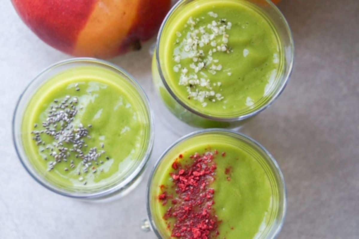 Tropical green smoothie in three glasses, each topped with either chia seeds, hemp hearts, or crushed freeze-dried strawberries. One mango to the back left.