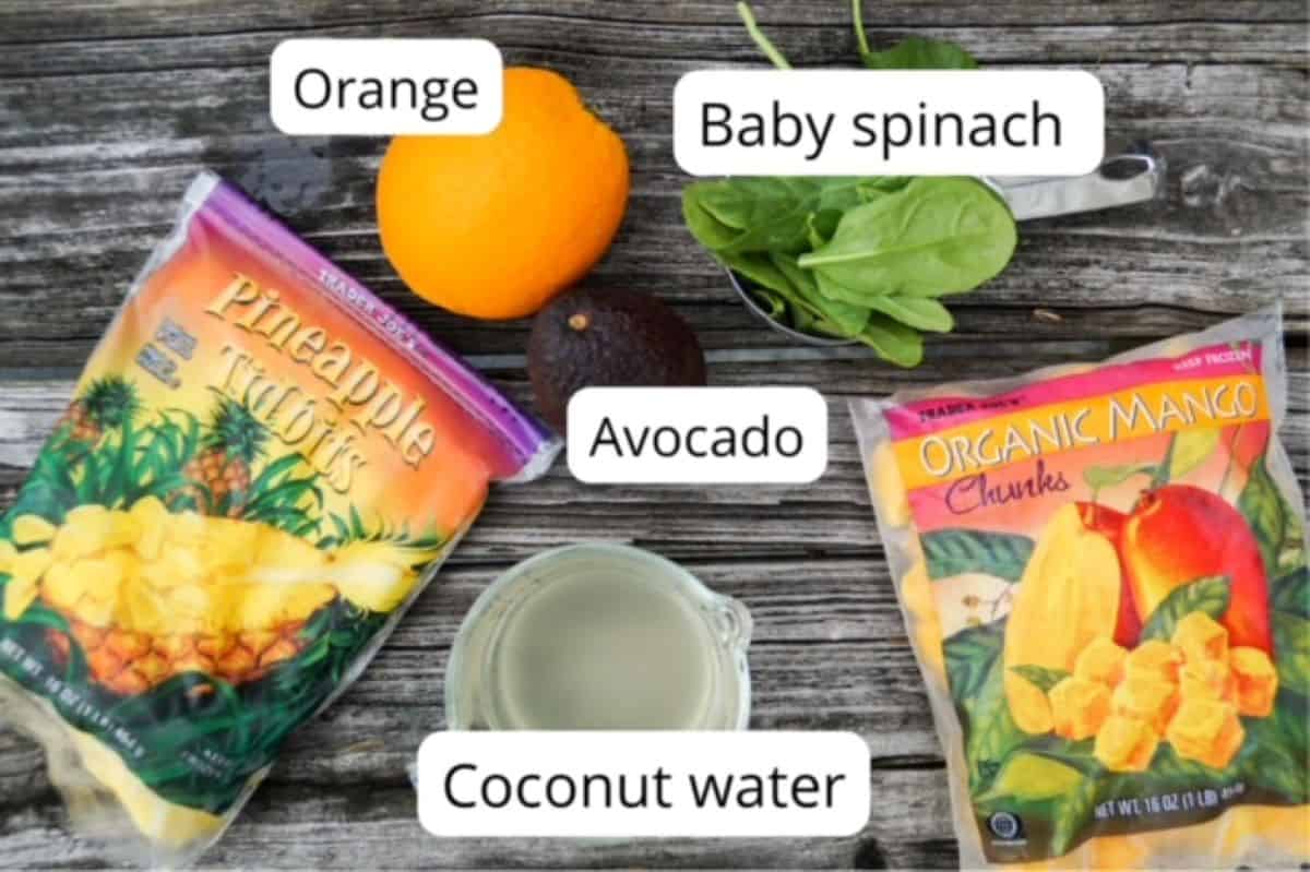 One bag of frozen pineapple pieces, one bag of frozen mango chunks, one naval orange, coconut water in a measuring cup, one avocado, and one overflowing cup of baby spinach pictured on a wooden background. All but the bags of frozen fruit are labeled.