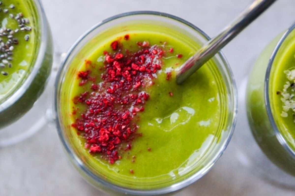 Up close image of the top of a glass with tropical green smoothie topped with crushed freeze-dried strawberries served with a stainless-steel straw. Part of an additional smoothie is on each side of the smoothie, depicting three glasses of a green smoothie. 