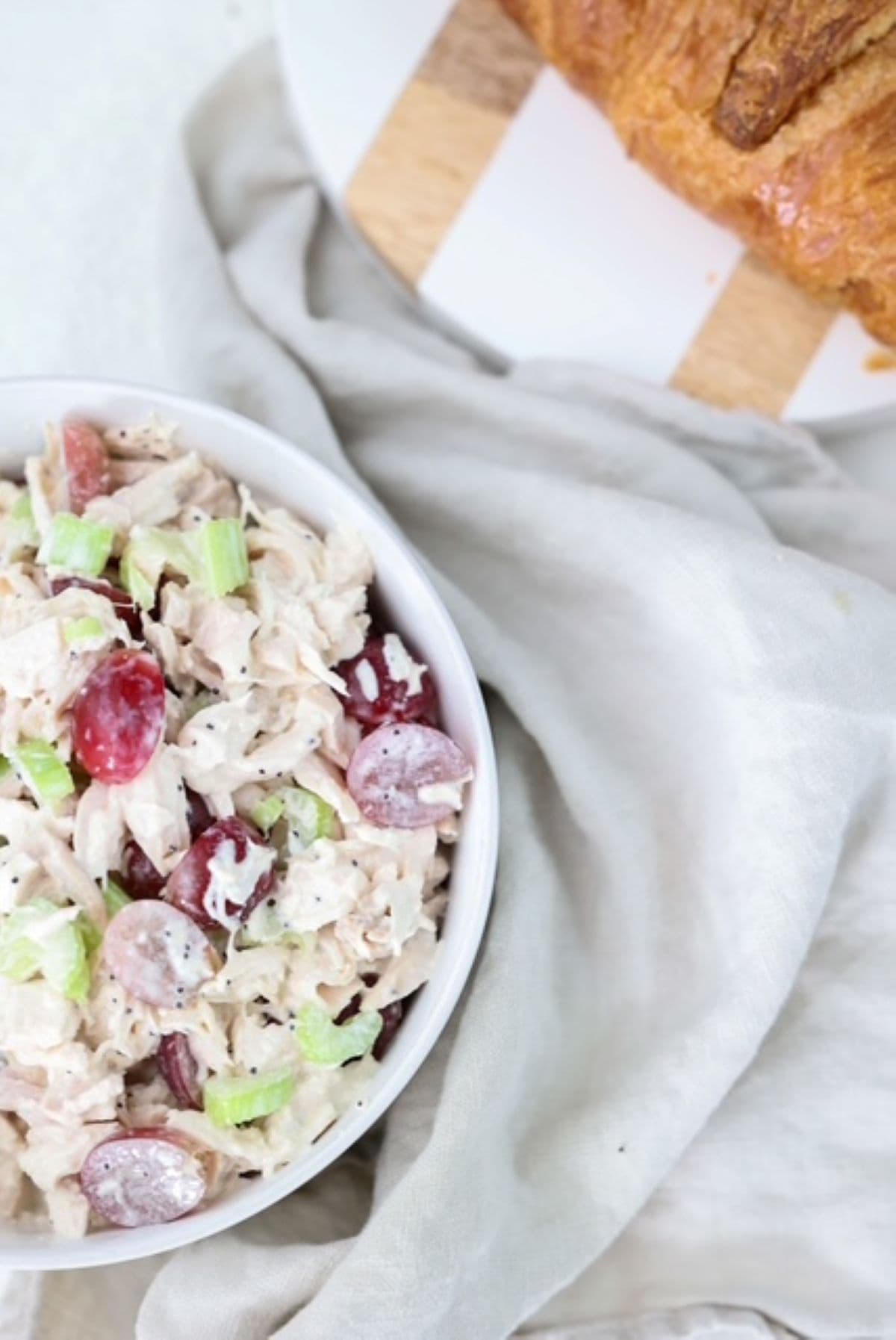 Chicken salad made with grapes and celery in a white bowl with a linen napkin and white and brown cutting board to the side.