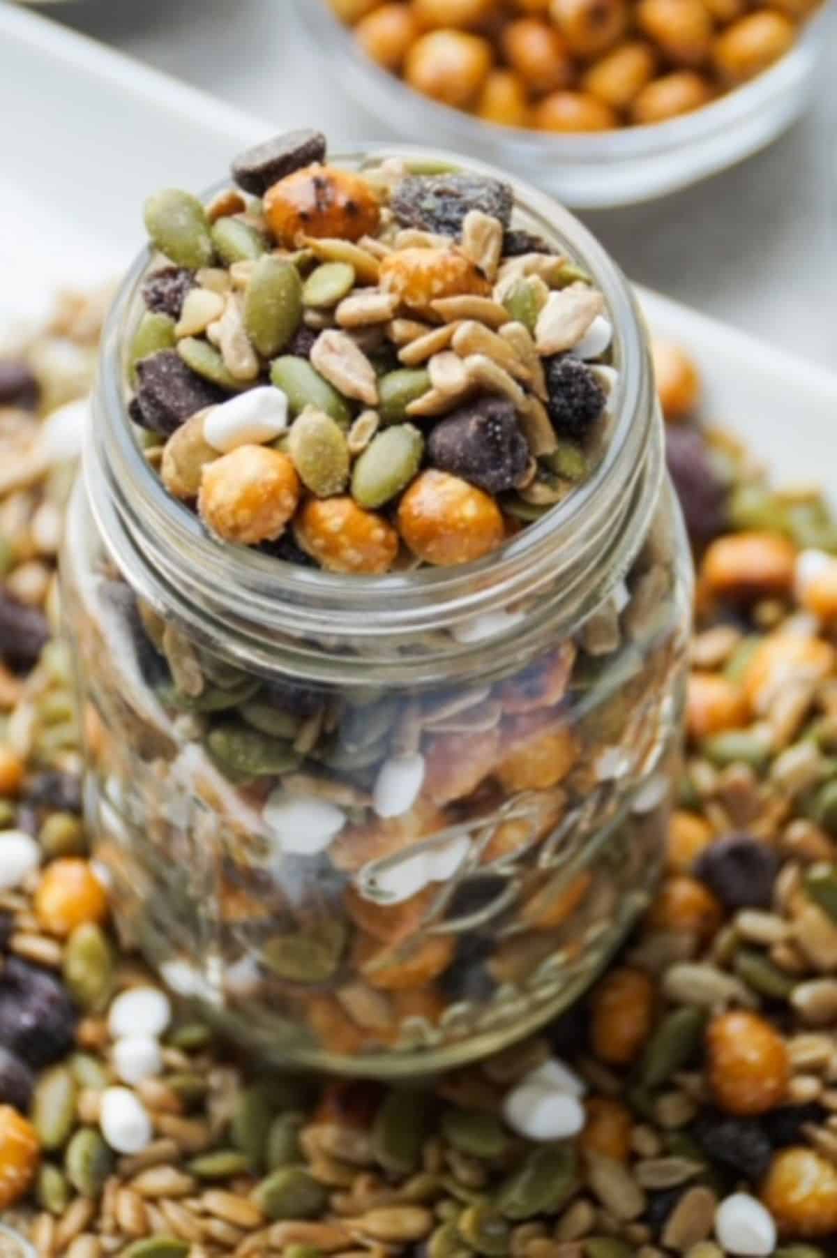 One glass mason jar filled with peanut-free trail mix. Base of jar surrounded by trail mix on a white plate. White bowl with pretzel balls partially pictured in the background.
