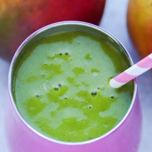 Up close image, top view of a green smoothie in a pink insulated wine tumbler with a pink and white straw. Two mangos are in the background.