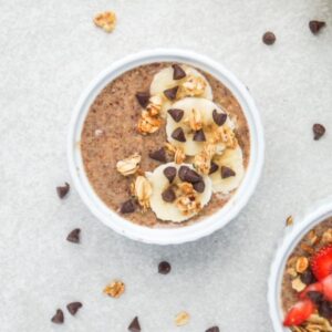 One serving of flaxseed pudding topped with banana slices, granola, and mini chocolate chips in the center, part of a second serving is showing in the bottom right corner, and mini chocolate chips and granola pieces are scattered around the surface.