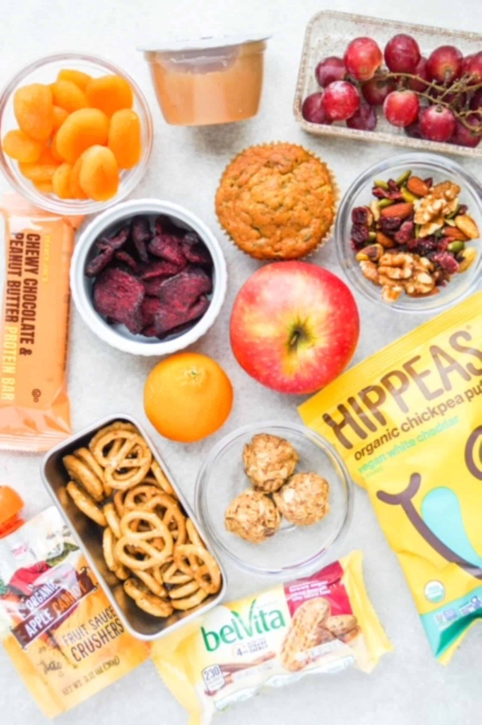 Individual snacks laid out on a surface: dried apricots, applesauce, grapes, beet chips, muffin, apple, trail mix, protein bar, spelt pretzels, HIPPEAS, energy bites, belVita package, and a apple-carrot fruit crusher from Trader Joe's.