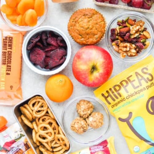 Lunch box snack ideas served individually: dried apricots, protein bar, beet chips, spelt pretzels, fruit crusher, energy bites, clementine, apple, bag of HIPPEAS, trail mix, and muffin.