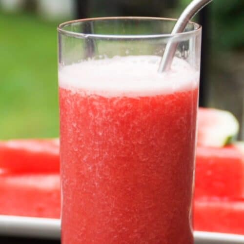 One tall glass of watermelon banana smoothie with a stainless-steel straw. A white plate with stacked watermelon slices is in the background.