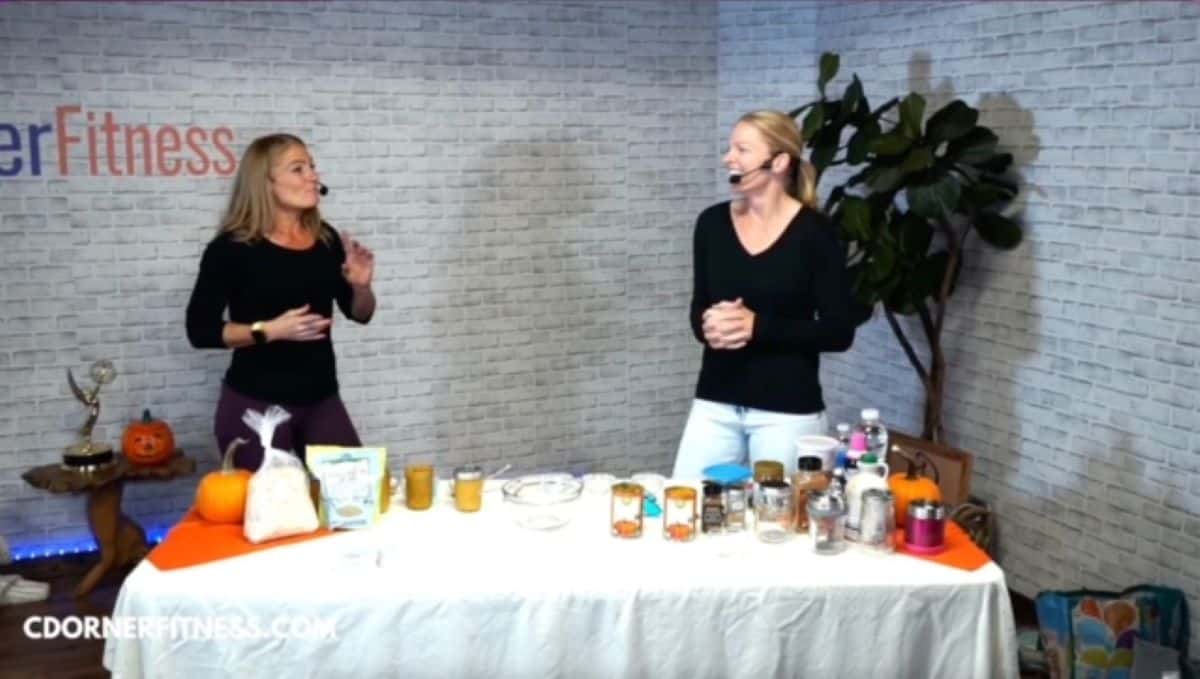 Chris Dorner and Kristi Ruth talking to each other while standing behind a table with a white tablecloth that has pumkin overnight oat ingredients as well as cooking utensils and food storage containers set up for a cooking demo. CDorner Fitness logo is on the back wall to the left. 